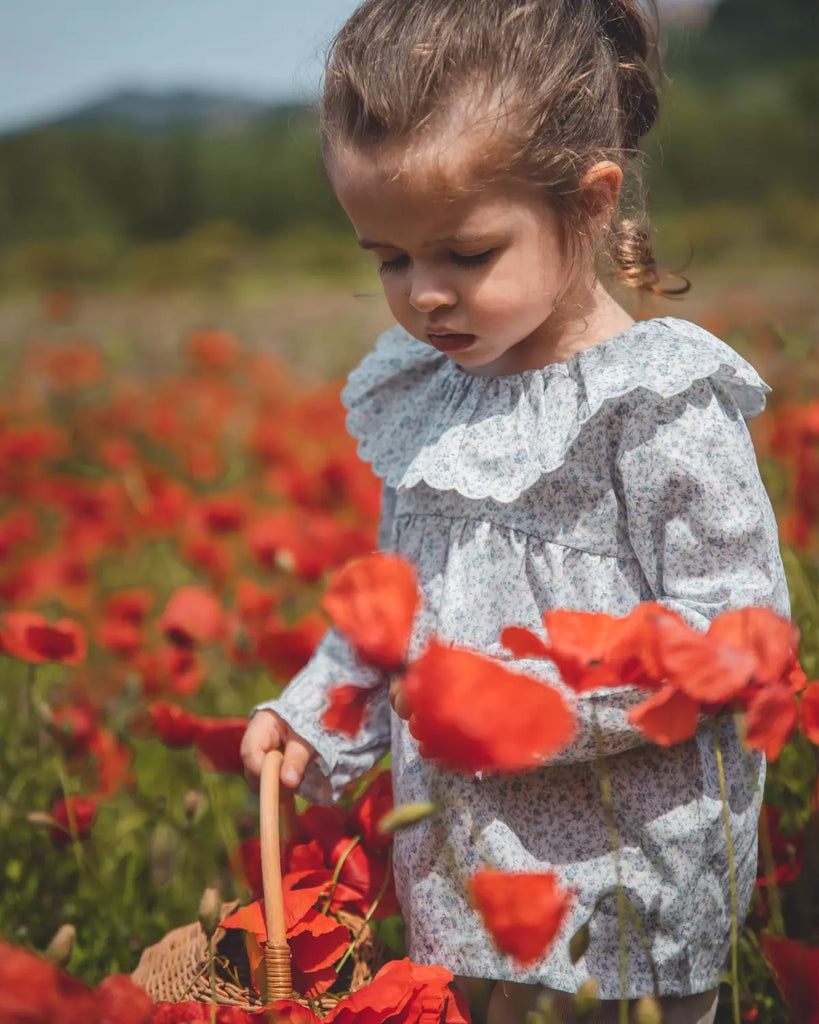 Image d'une petite fille dans un champ de cocquelicot, portant la blouse pour bébé fille à col volanté et fleurs bleues de la marque Bobine Paris.