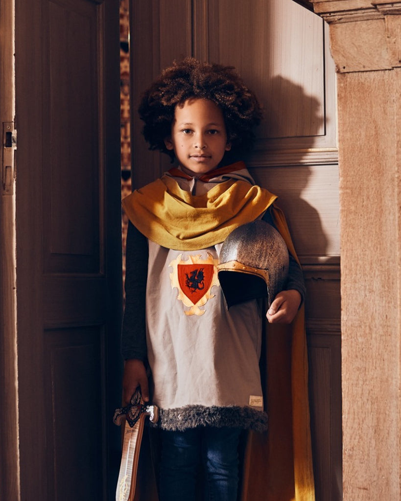 Petit garçon portant une tunique de chevalier, une cape jaune, un casque de chevalier et une épée.