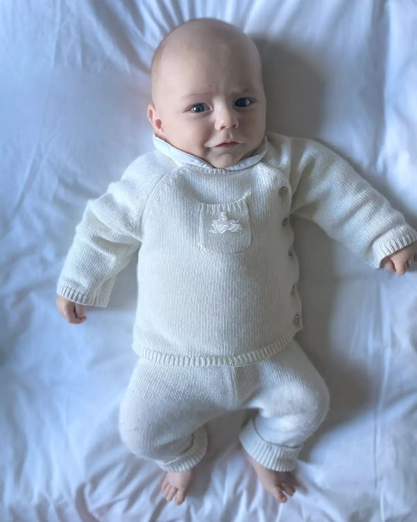 Image d'un bébé portant le pull bébé cache-coeur avec pompon écru en laine et cachemire de la marque Bobine avec un pantalon assorti.
