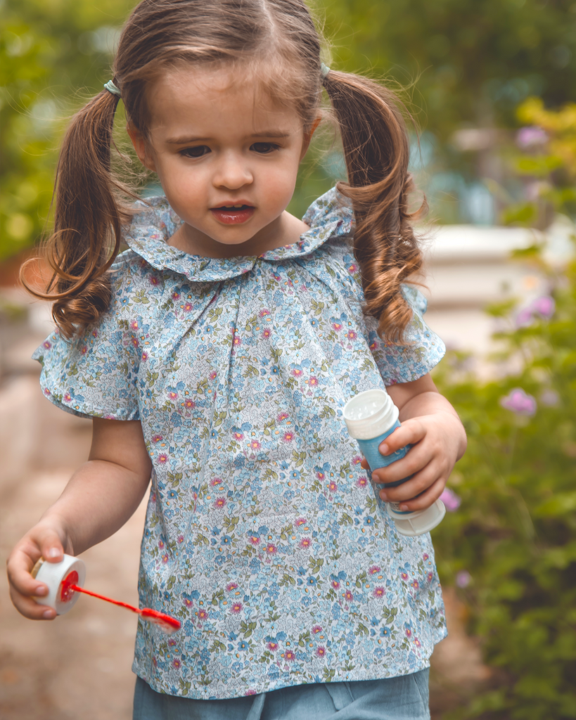 Photo d'une petite fille portant la blouse à col volanté pour bébé fille à motifs fleuris bleus de la marque Bobine Paris pendant qu'elle joue avec des bulles..