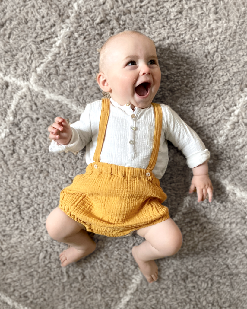 Bloomer pour bébé à bretelles en gaze de coton couleur curry de la marque Bobine Paris porté avec un chemisier blanc en gaze de coton.