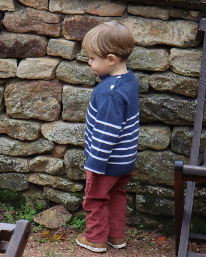 Petit garçon portant un haut manches longues bleu à rayures blanches et un pantalon à velours côtelé rouge.