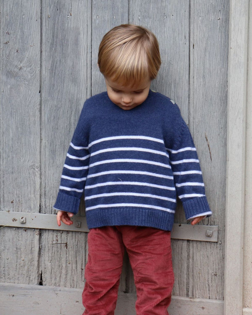 Petit garçon portant un haut manches longues bleu à rayures blanches et un pantalon à velours côtelé rouge.