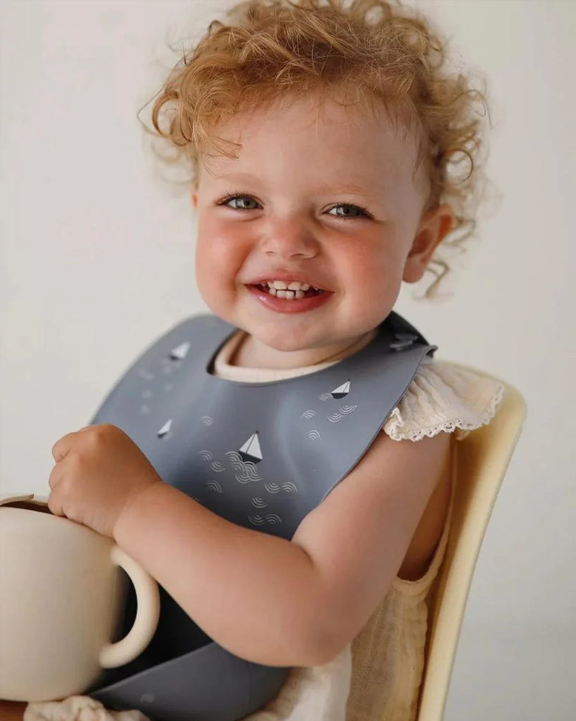 Petite fille souriant en portant le bavoir en silicone gris à motifs bateaux de la marque Bobine Paris.