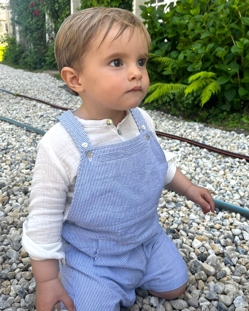 Léger zoom du look porté composé d'une salopette courte à rayures bleues et d'une chemise en gaze de coton retroussée aux manches pour bébé garçon.