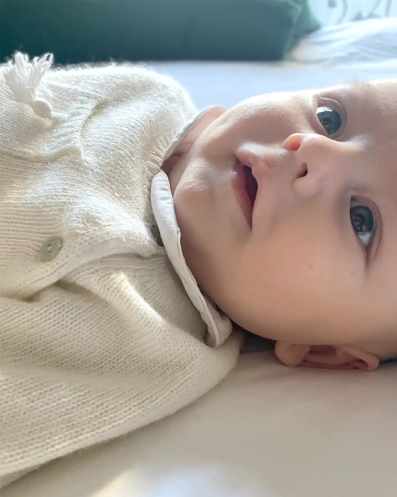 Image d'un bébé portant le pull bébé cache-coeur avec pompon écru en laine et cachemire de la marque Bobine avec une chemise blanche.