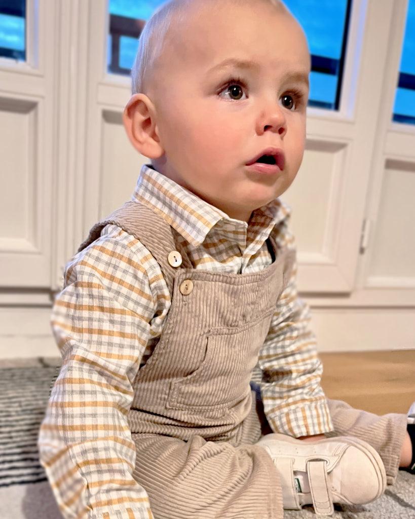Bébé portant une salopette beige sable en velours avec en dessous une chemise à carreaux beiges et gris