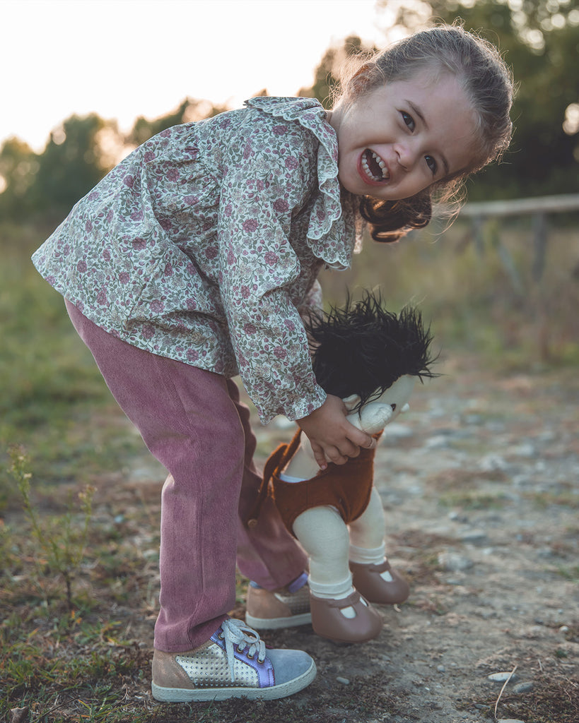 Petite fille portant une blouse à imprimés mauves doté d'un col volant et un pantalon rose tenant dans ses mains un doudou en forme d'humain.