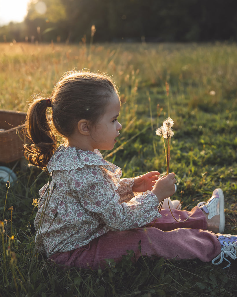 Petite fille assise dans un pré portant une blouse à imprimés mauves doté d'un col volant et un pantalon rose tenant dans sa main des pissenlits.