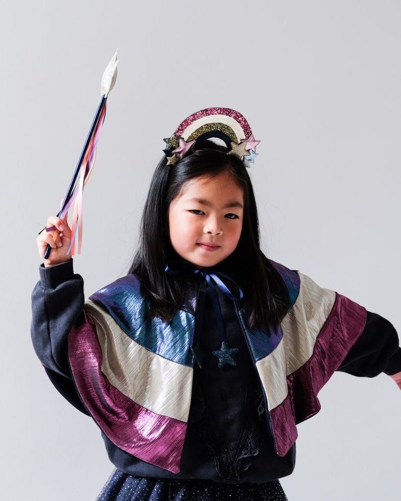 Petite fille portant une cape courte arc-en-ciel avec un serre-tête et une baguette magique assortis