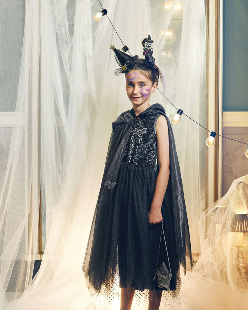 Petite fille portant une cape en tulle noire, ornée d'empiècements chauve-souris et d'une capuche avec sa robe, son sac et son serre-tête assortis.